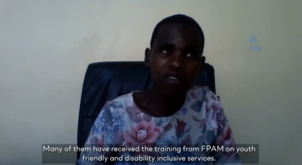 Elizabeth Kanjiwa, a young Malawian woman with vision impairments, has short black hair. She is seated inside a room in front of a white wall. She is communicating with Braille to present her "Global Disability Summit - Spotlight on Gender" speech. She wearing a pink, blue, white, and purple blouse with flower designs.