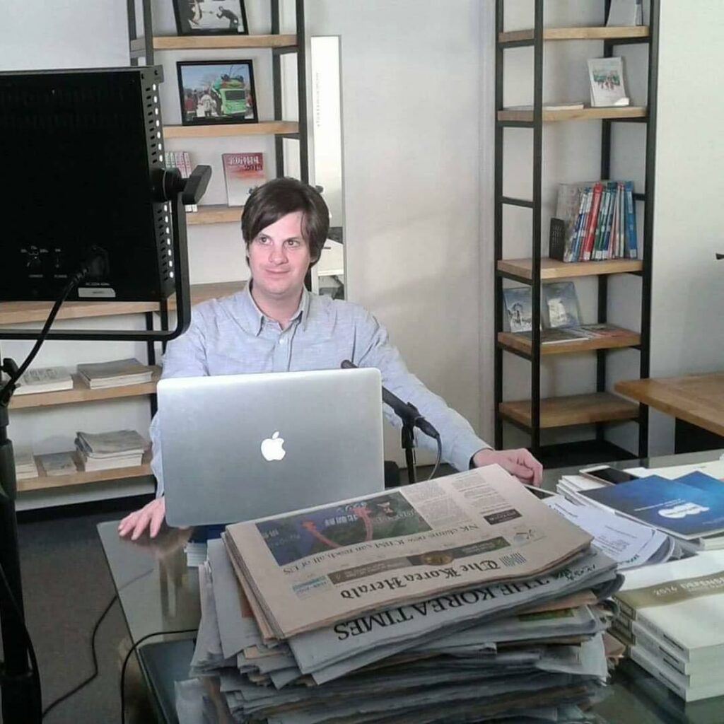 Jason Strother, a white, middle-aged man with short brown hair, sits a desk stacked with various newspapers and a MacBook laptop. He is wearing a gray button-down shirt.
