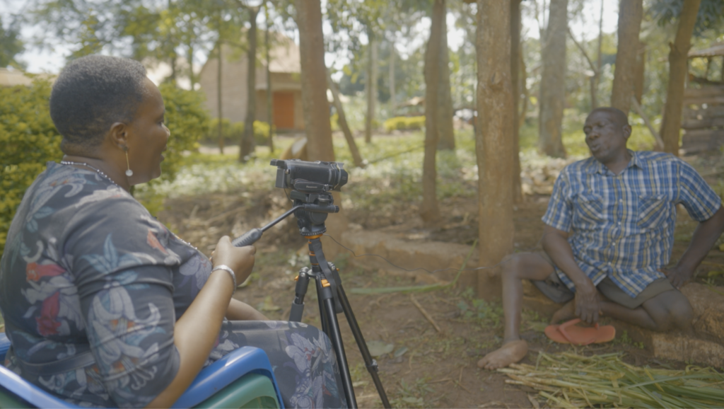 DJP Fellow Christine Dhikusooka interviews Steven Bukaya at his farm in Jinja. She is holding the camera, and Steven is sitting on the ground smiling at her. 