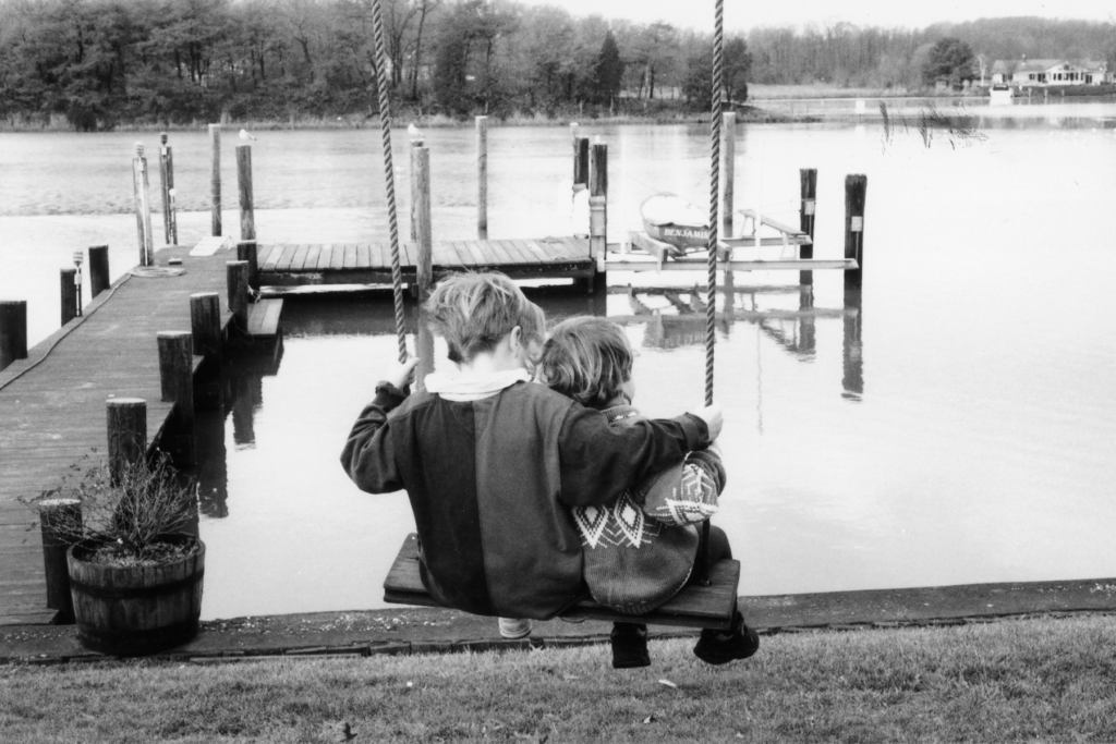 A black-and-white photo of Marian and David from behind. They are children, and David has his arm around Marian as they sit on a swing and look out at the water.