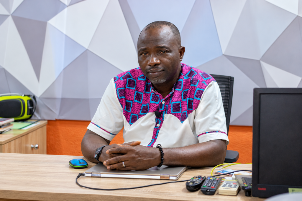 Afolabi Fajemilo, co-founder of the Festus Fajemilo Foundation in Nigeria, sits behind a desk wearing a white shirt with bright pink and turquoise geometric patterns.