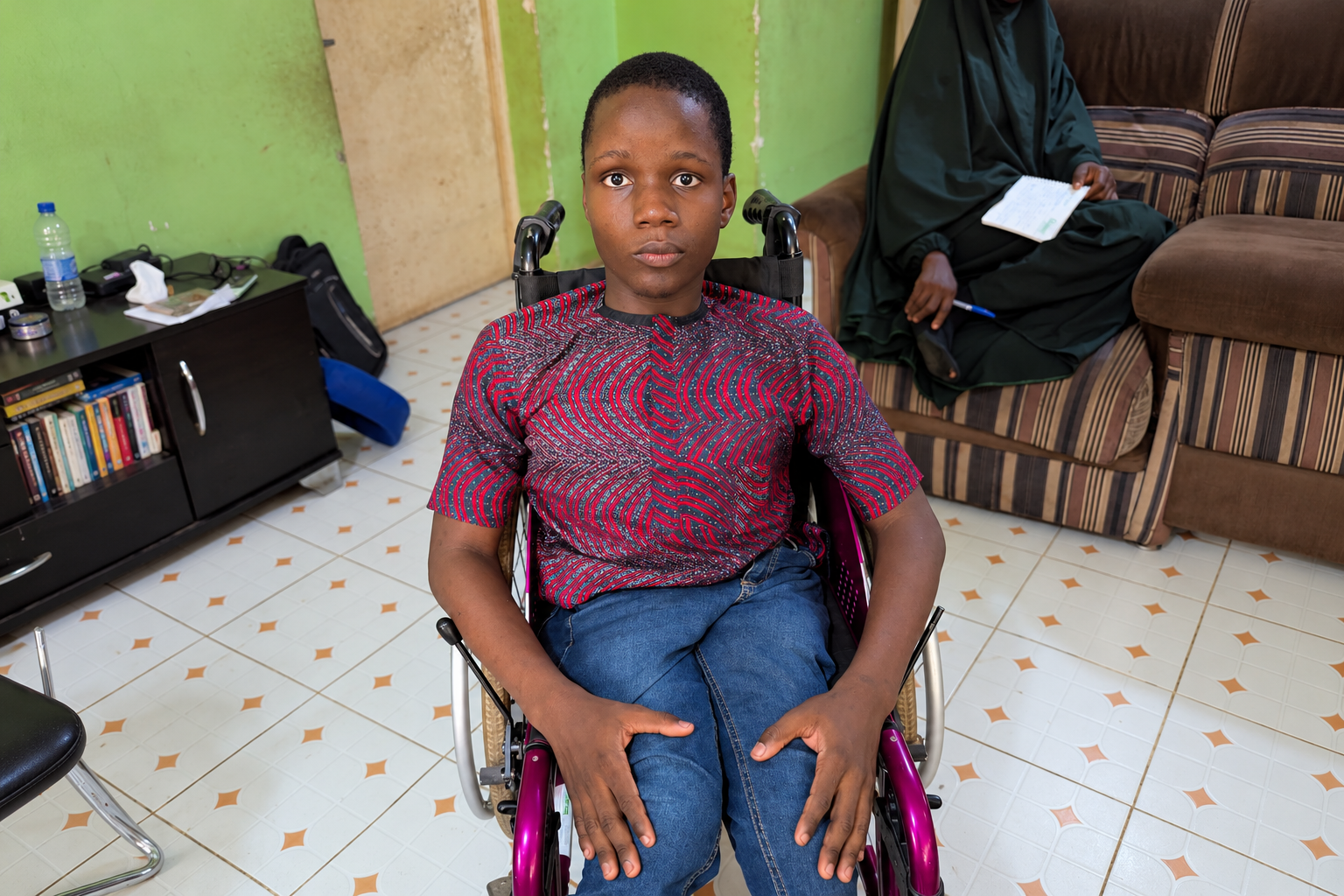 Festus Fajemilo, a young Nigerian wheelchair user wearing a red-and-black patterned shirt and blue jeans, looks directly at the camera while seated in a room with green walls.