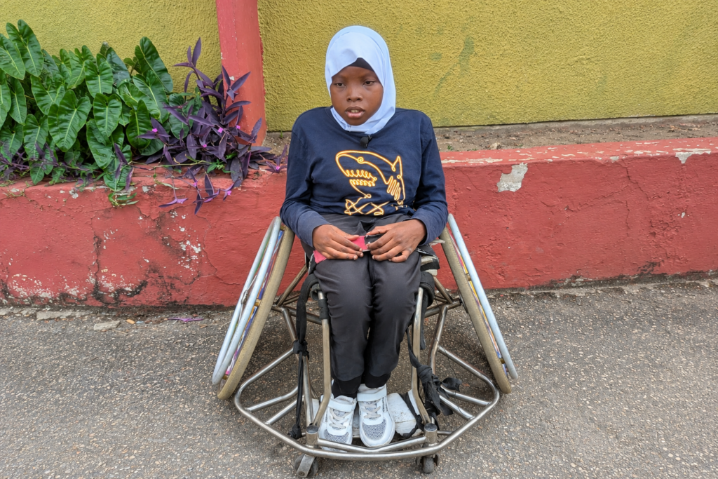 Samara, a young Nigerian girl wearing a light blue hijab, a navy sweatshirt with a yellow shark design and black pants, sits in a wheelchair outdoors.