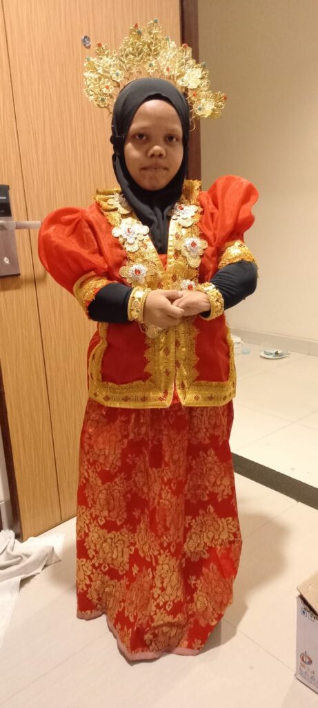 Dija, an Indigenous Indonesian woman with a disability, poses for the camera. She is holding her own hands. She is wearing bright orange Bodo (Bugis-Makassar) clothes. She is wearing a black hijab with a headband (pinang goyang) on her head, a multi-layered necklace, and kroncong bracelets on both wrists, all of which are gold. 