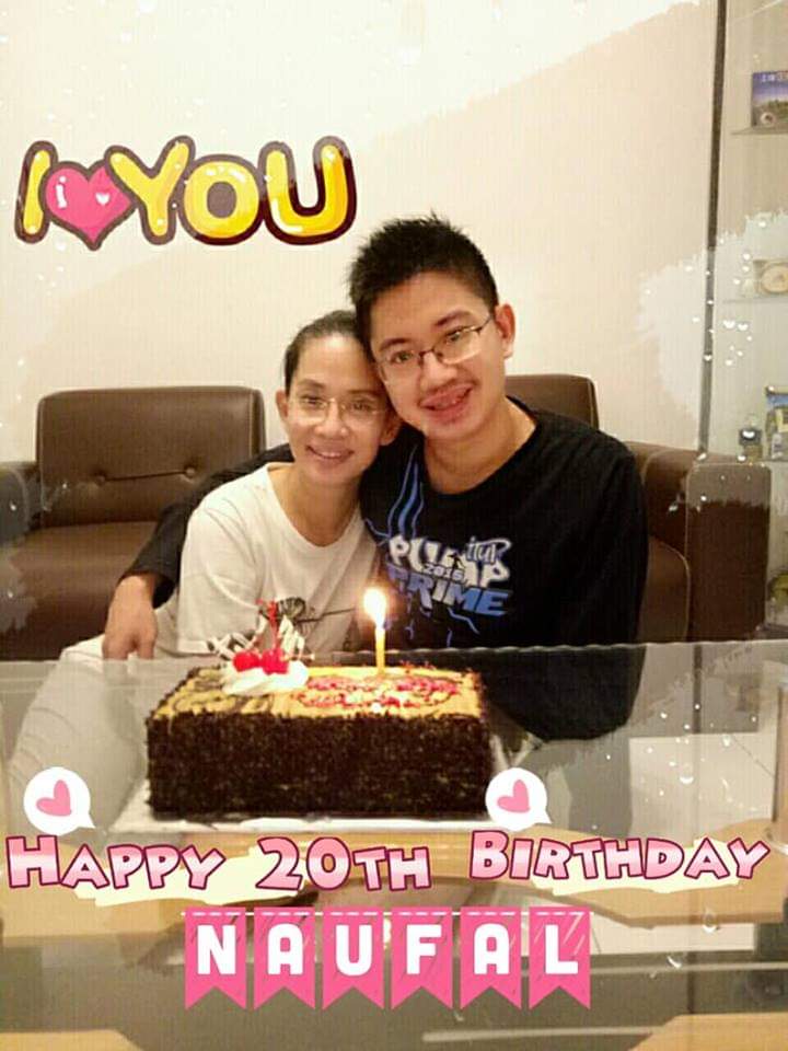 Naufal Asy-Syaddad, a young Indonesian man with autism, poses with his mother, Lani Setyadi, an Indonesian woman, for his 20th birthday. Naufal is wearing glasses and a black shirt with blue and white words and designs. His hand is over his mother's shoulders; she is seated to the left. Setyadi is wearing glasses and a white shirt and jeans. There are two brown sofa chairs behind them. A brown and beige cake with whipped cream, two cherries, and a lit candle at the top rests on a glass table in front of Asy-Syaddad and Setyadi. Pink and white text at the bottom of the image says, "HAPPY 20TH BIRTHDAY NAUFAUL." At the top of the image, text says "I" in yellow, followed by a a pink heart icon and text that says "YOU" in yellow.