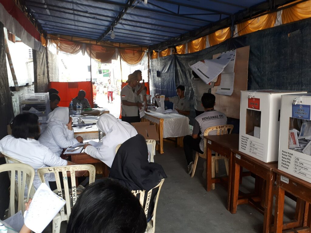 Eleven Indonesian people are seated under a tent with brown tables and white chairs. Two seated women are wearing white hijabs and white shirts. Some seated men are wearing grey shirts with black designs. Two other men are standing next to each other on the right, and one speaks into a microphone. A third man standing up looks through large sheets of paper hung on the tent’s wall on the right. Some seated people are looking at the two men standing up. On the right, two boxes rest on a table.