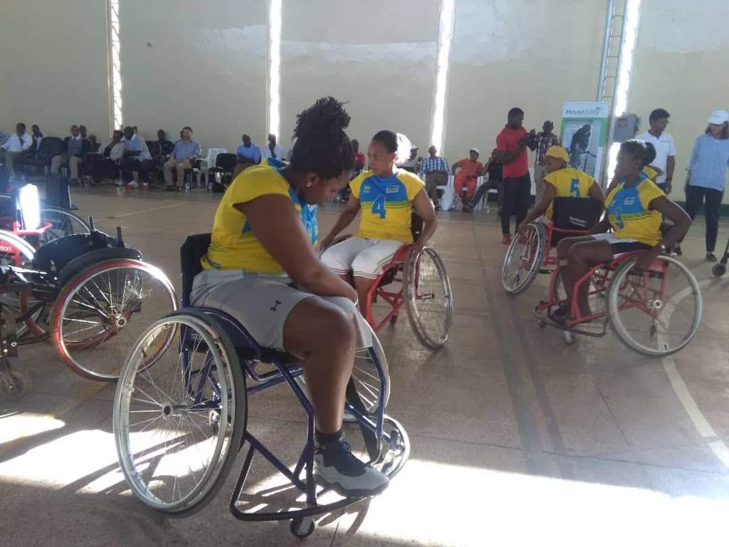 Mukangoga adjusts her multisport wheelchair. 3 women wearing yellow jerseys push their wheelchairs behind  her.
