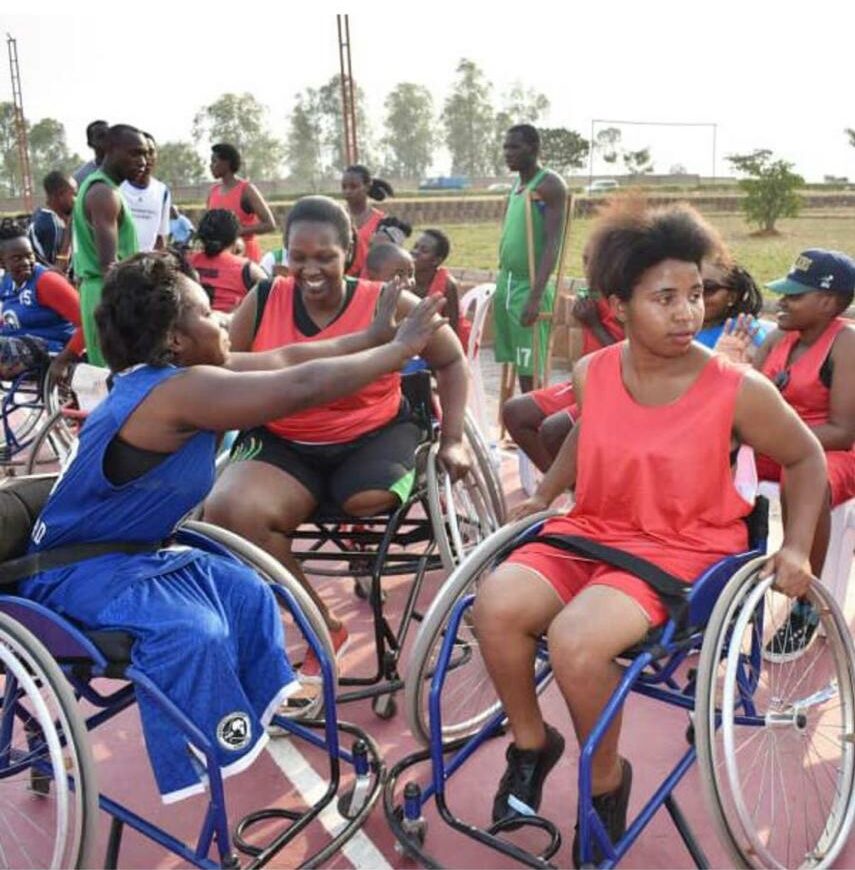Three women in red and blue uniforms play wheelchair basketball on a crowded court.