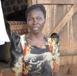 Rose Namulondo, an older Ugandan woman, sits in front of a wooden house. Hair hair is up, and she's wearing a black, yellow, and orange patterned dress.