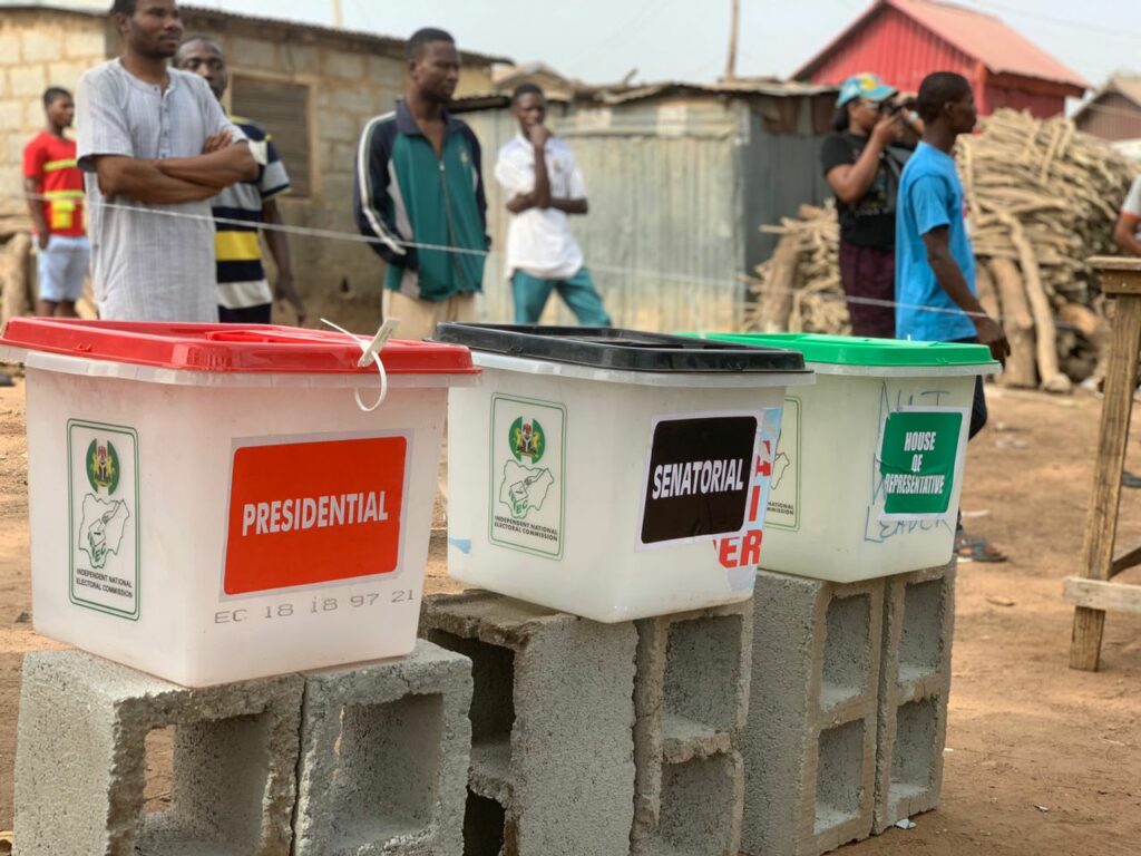 Three white bins are placed on concrete blocks. One says "presidential," while the second ssays "senatorial." The third says "House of Representatives." People are standing in line on a dirt road behind them.