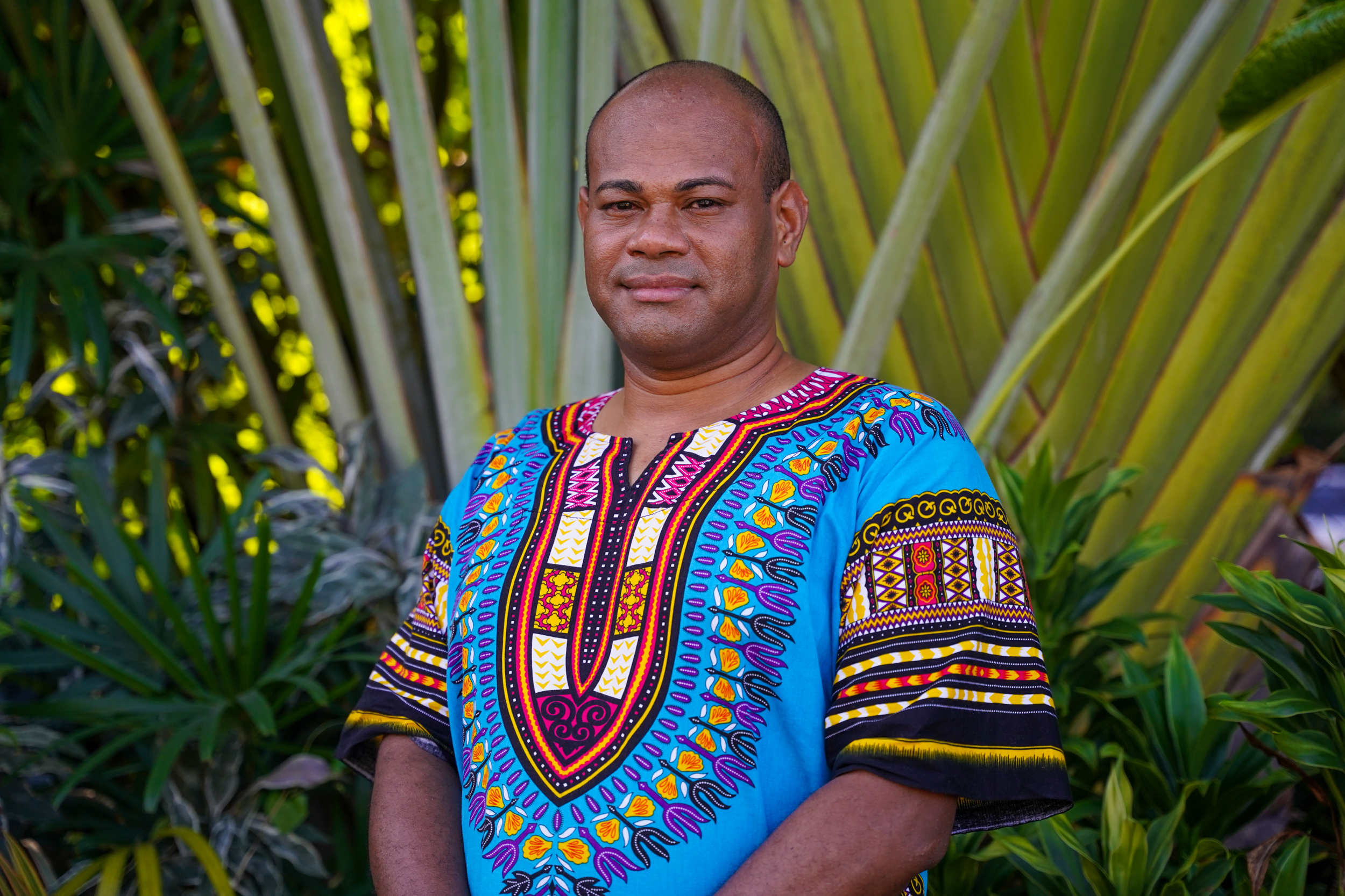 Isoa Nabainivalu stands outside and looks straight at the camera. A palm tree is behind him. He is wearing a traditional Fijian tunic shirt.