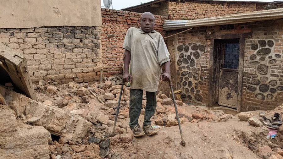 Thacien Nzigiyimana stands in front of rubble outside one-story stone buildings.
