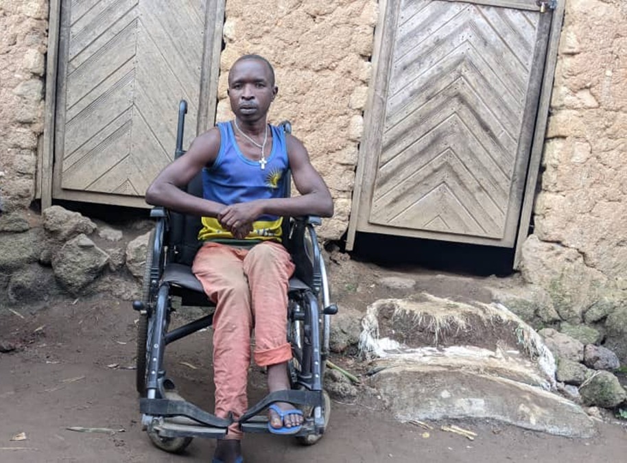 Theophile Nsengimana sits in a wheelchair in front of a stone building with two wooden doors.