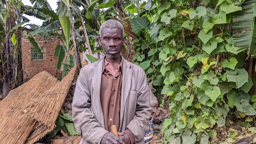 Bigirwanabagabo Kabagwera stands in front of a collapsed thatched roof.