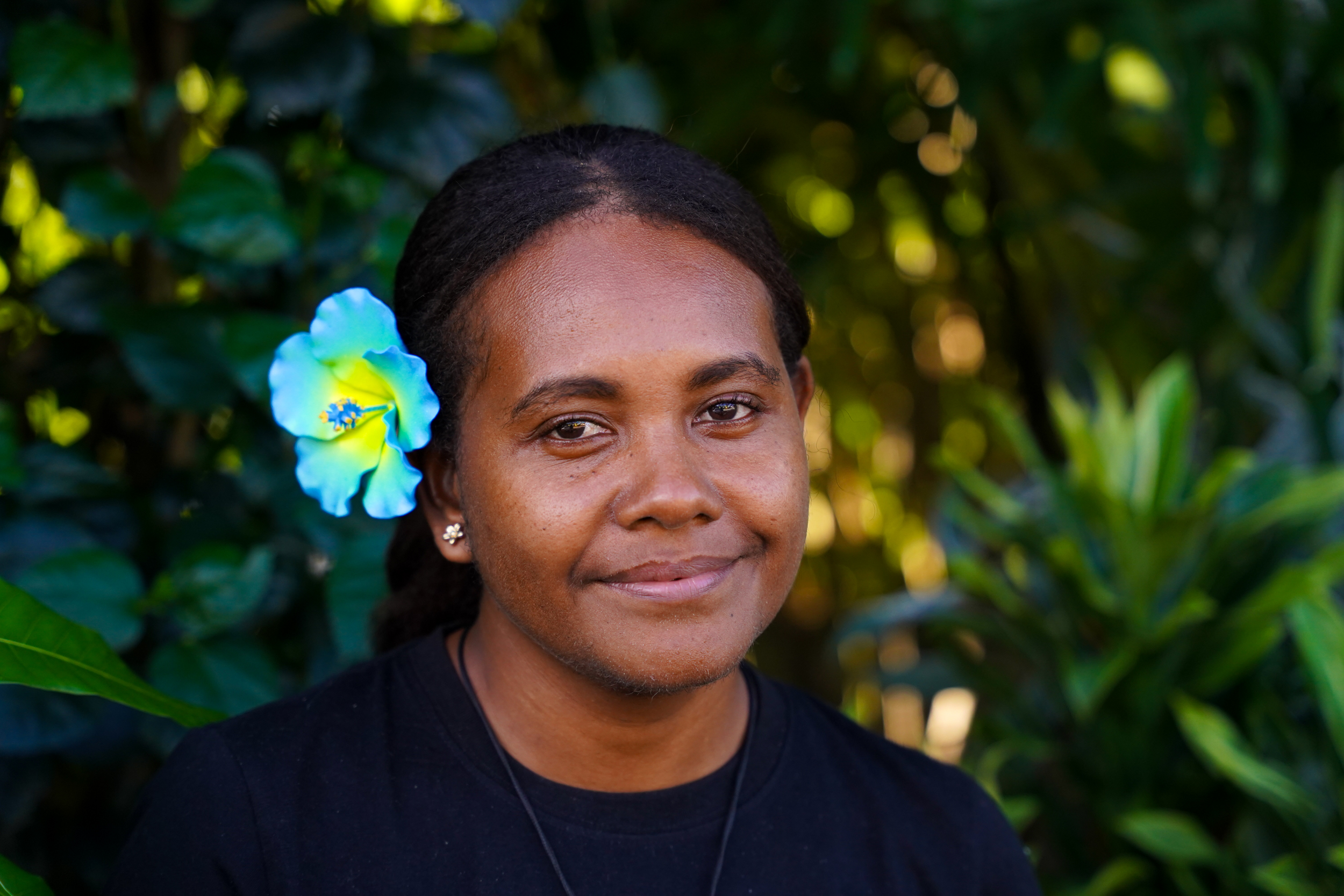 Melvina Voua smiles at the camera outside. She is wearing a flower in her hair, and green shrubs are behind her.