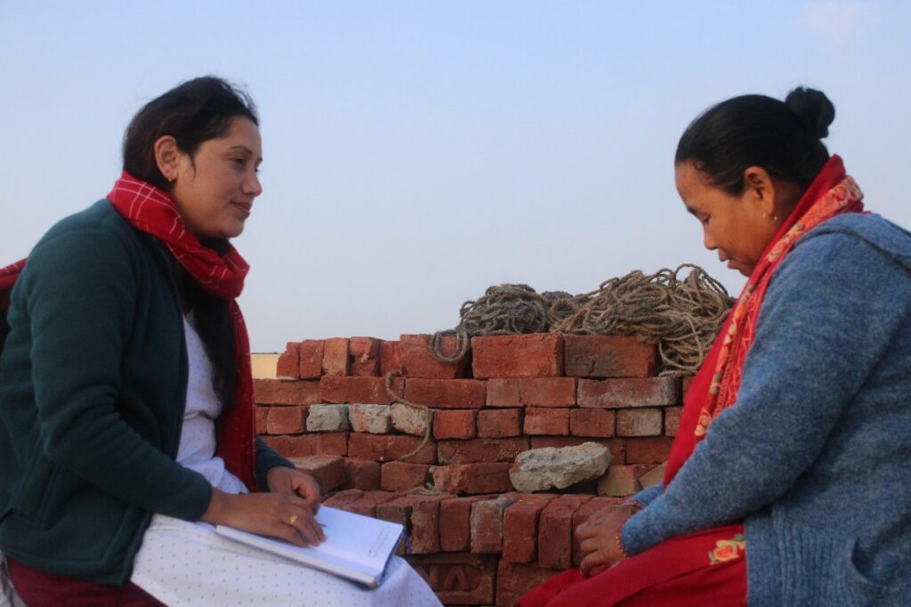 Srijana sits and talks with a Nepali woman. She has a notebook open on her lap.