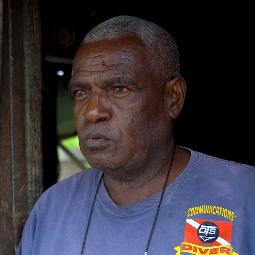 A ni-Vanuatu man leans on the wall of a shed and looks off into the distance.