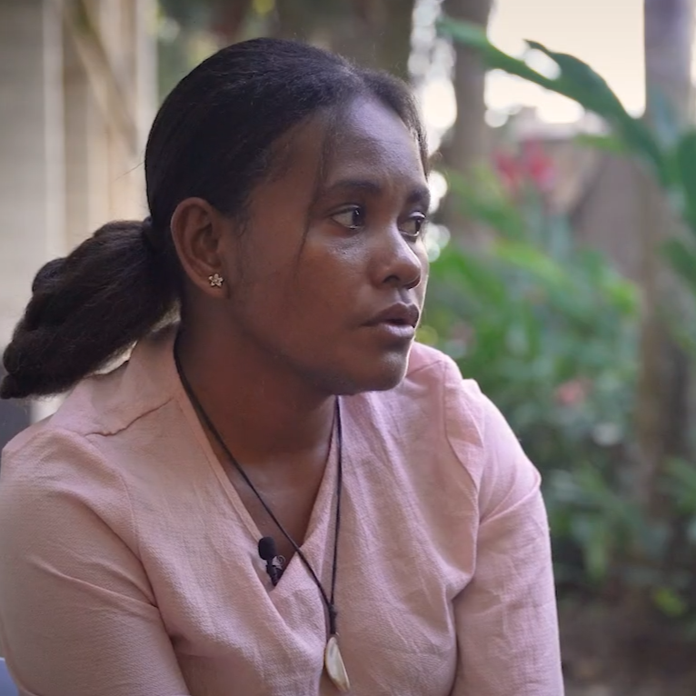 A young, dark-skinned woman from the Solomon Islands sits outside and looks off into the distance.