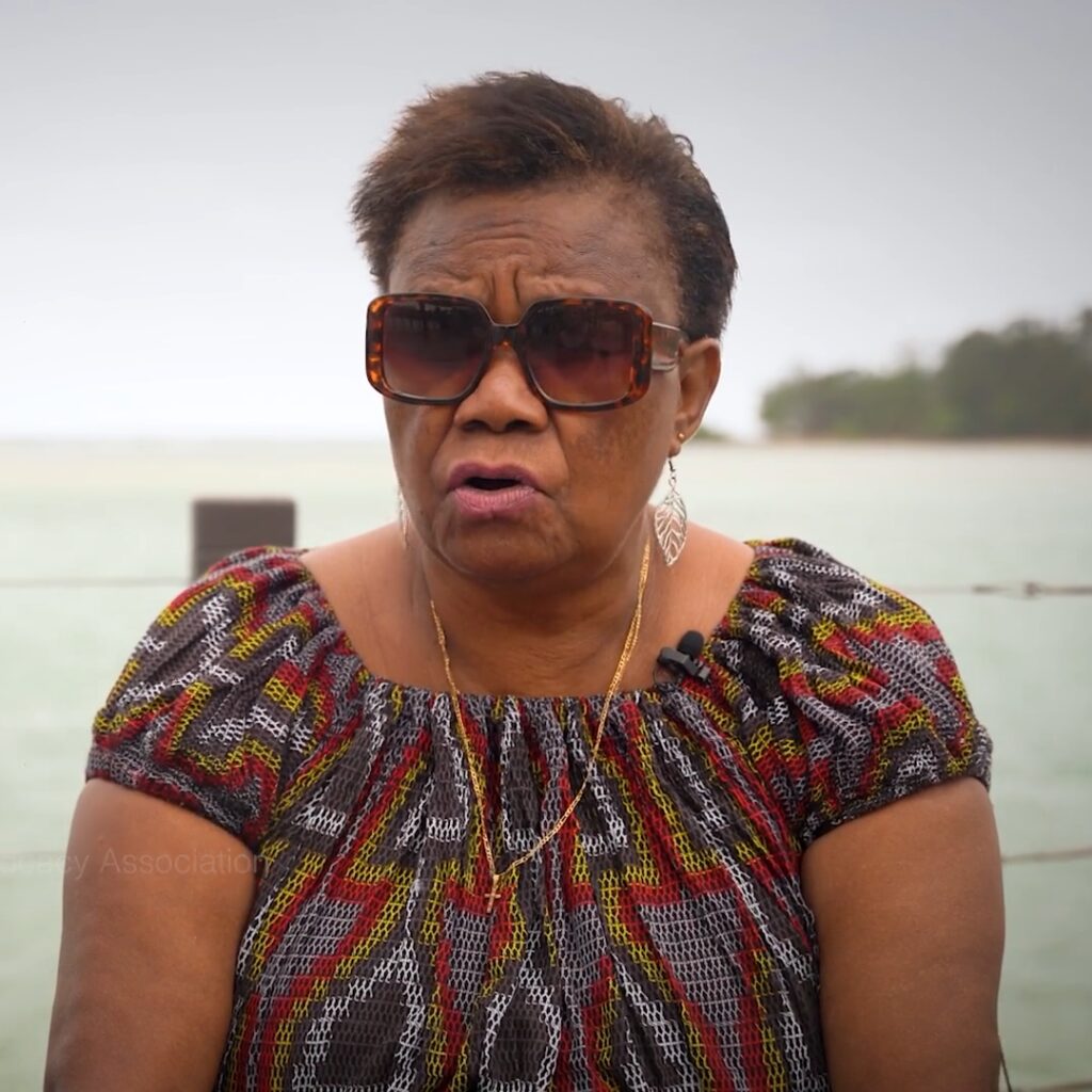 A ni-Vanuatu woman sits outside, with the ocean behind her, speaking to camera.