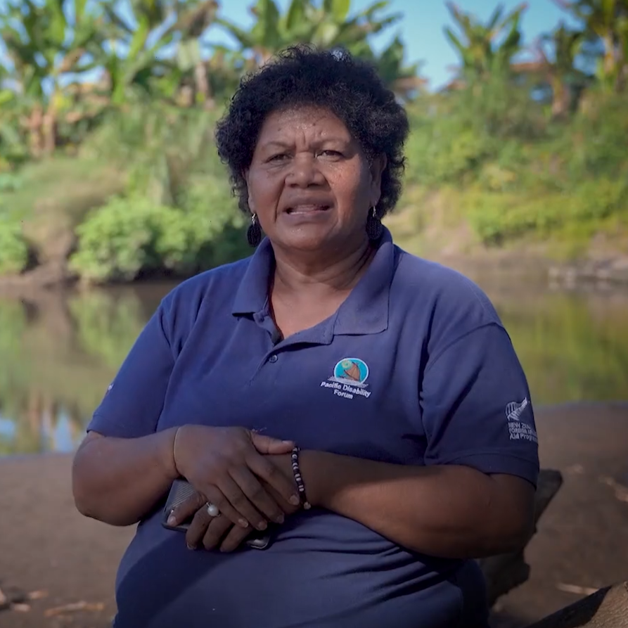 A Fijian woman sits on a branch outside and speaks to camera.