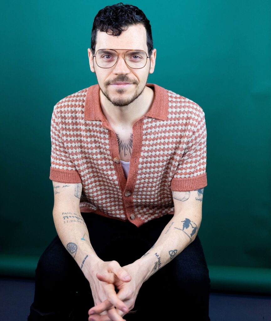 Dom Kelly smiles into the camera. He is a white man with glasses and a goatee. He has tattoos on his chest and arms.