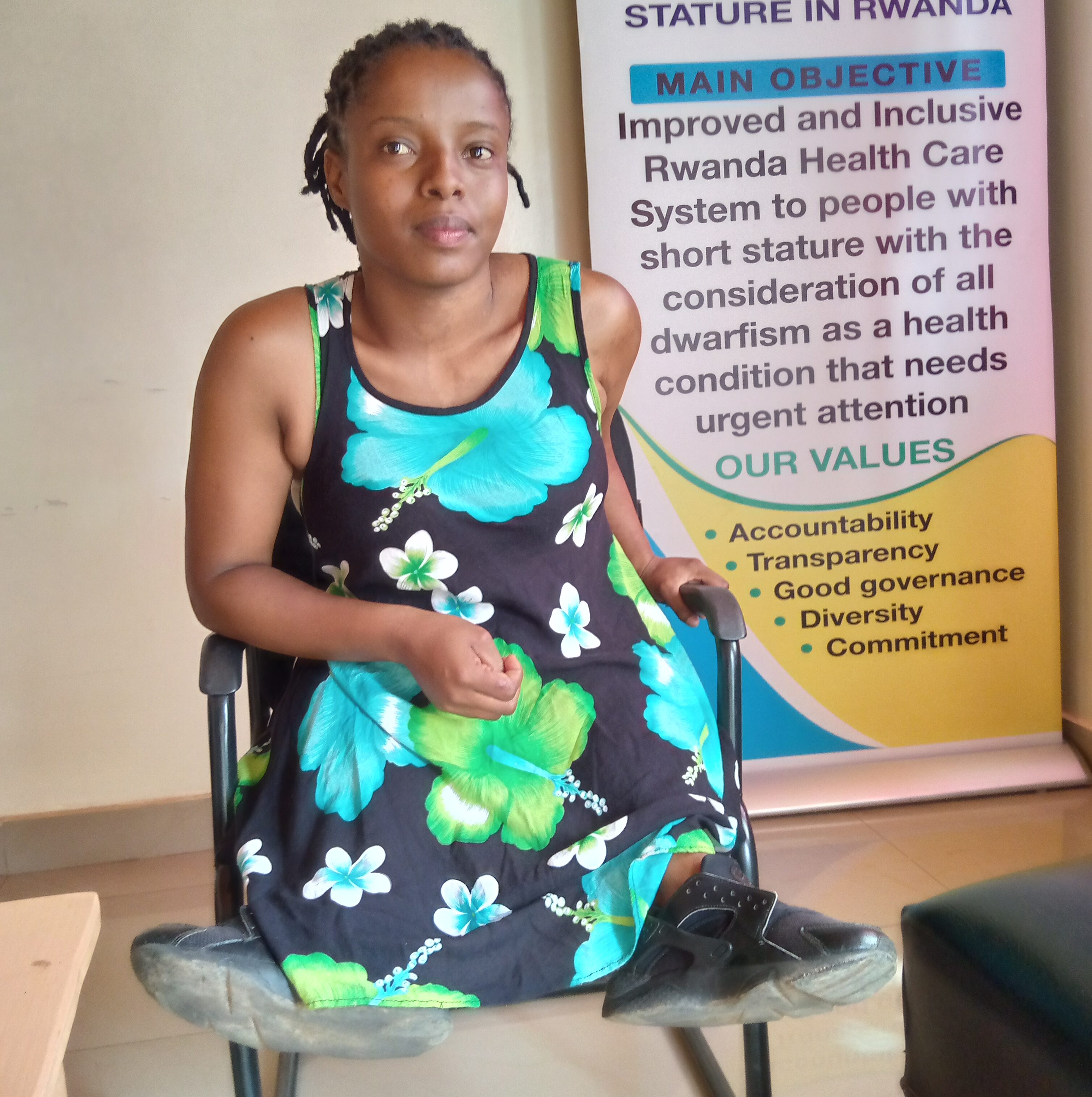 Alliance Ukwishaka, a young Rwandan woman, sits in a wheelchair in front of a banner about inclusive health services.