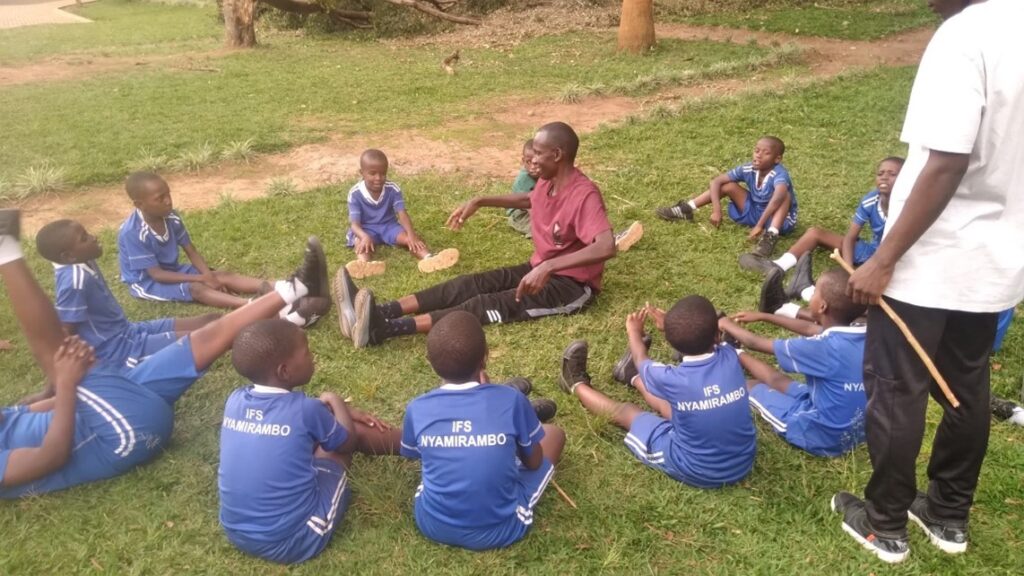 Jean Marie Furaha sits on the ground, surrounded by his young students wearing sports uniforms.