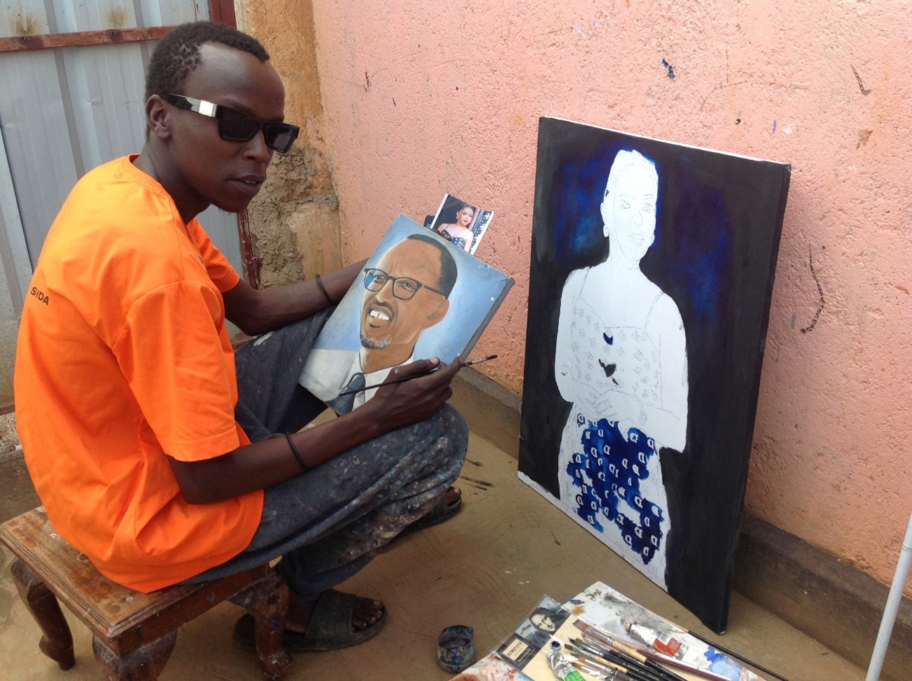 Jean de Dieu Uwikunda kneels next to one of his paintings.