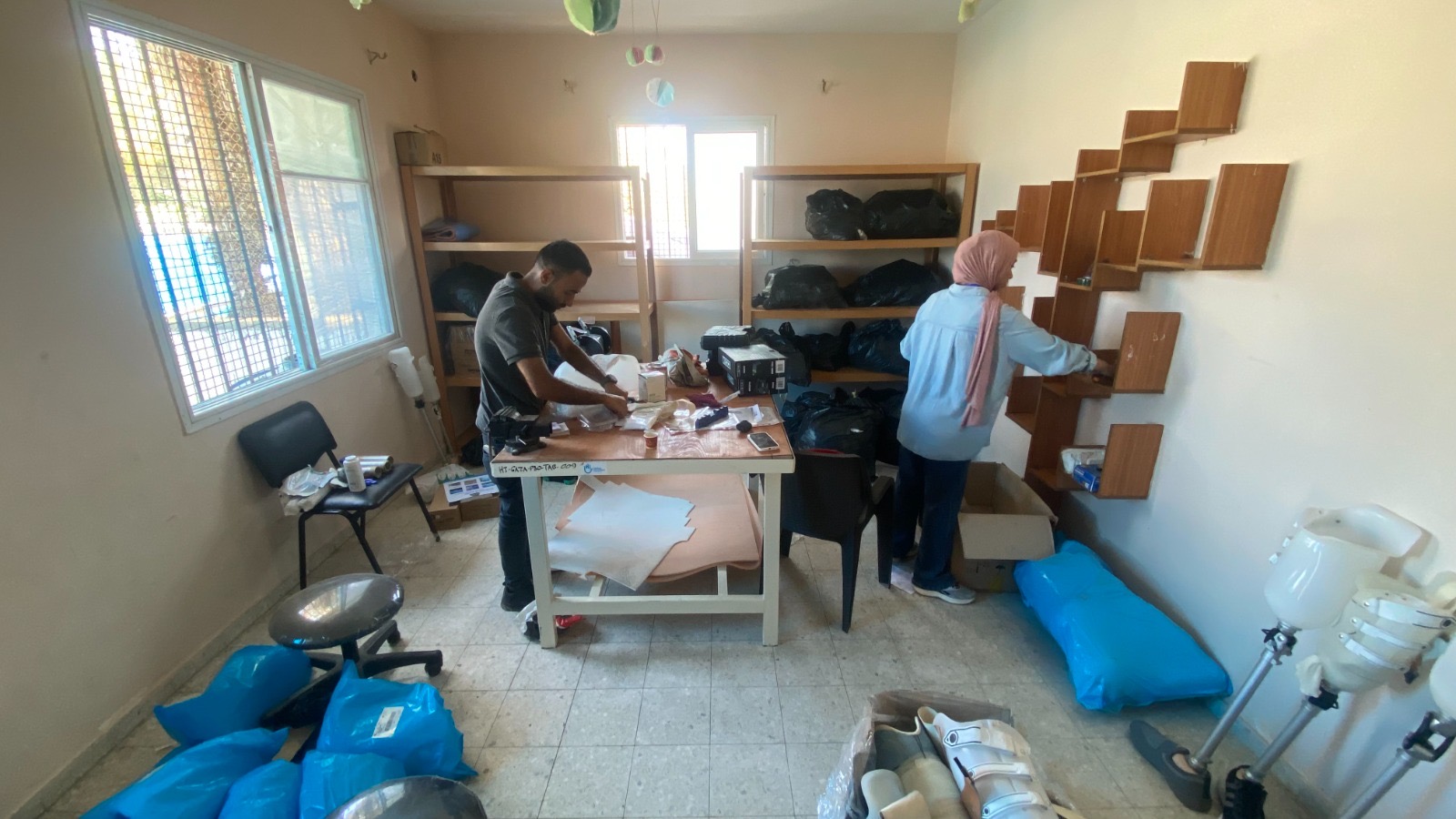 Two staff members at the Nahla Prosthetics & Orthotics Center in Gaza work in a small room, organizing medical supplies and prosthetic components.