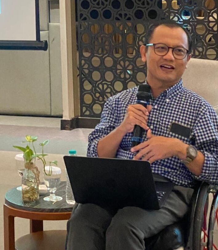 Bahrul Fuad, wearing glasses and shirt, sitting in his wheelchair while speaking into a microphone with a projector screen behind him.