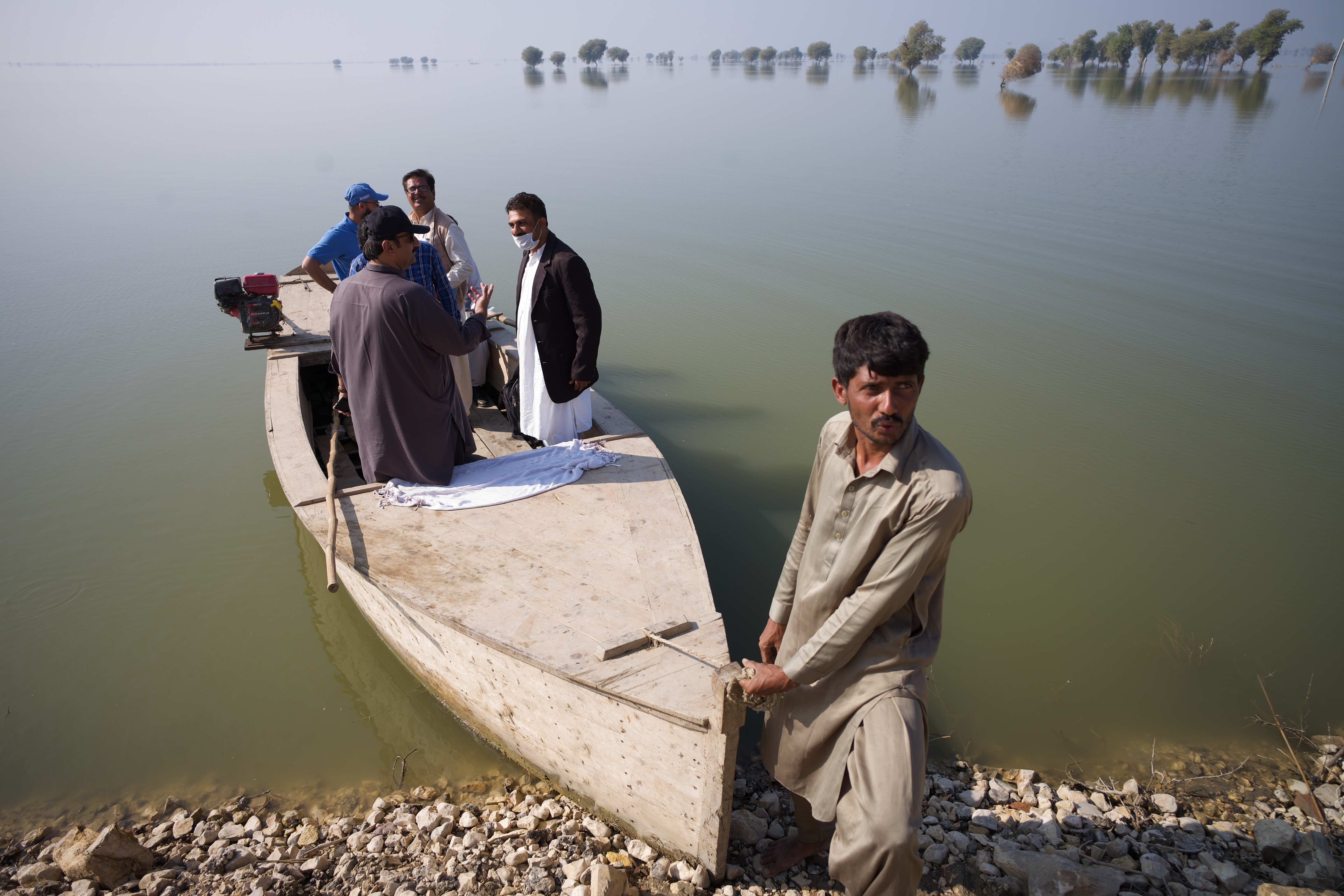A man pulls a wooden boat to shore. Four men are standing in the boat. Behind them, flood waters stretch to the horizon.