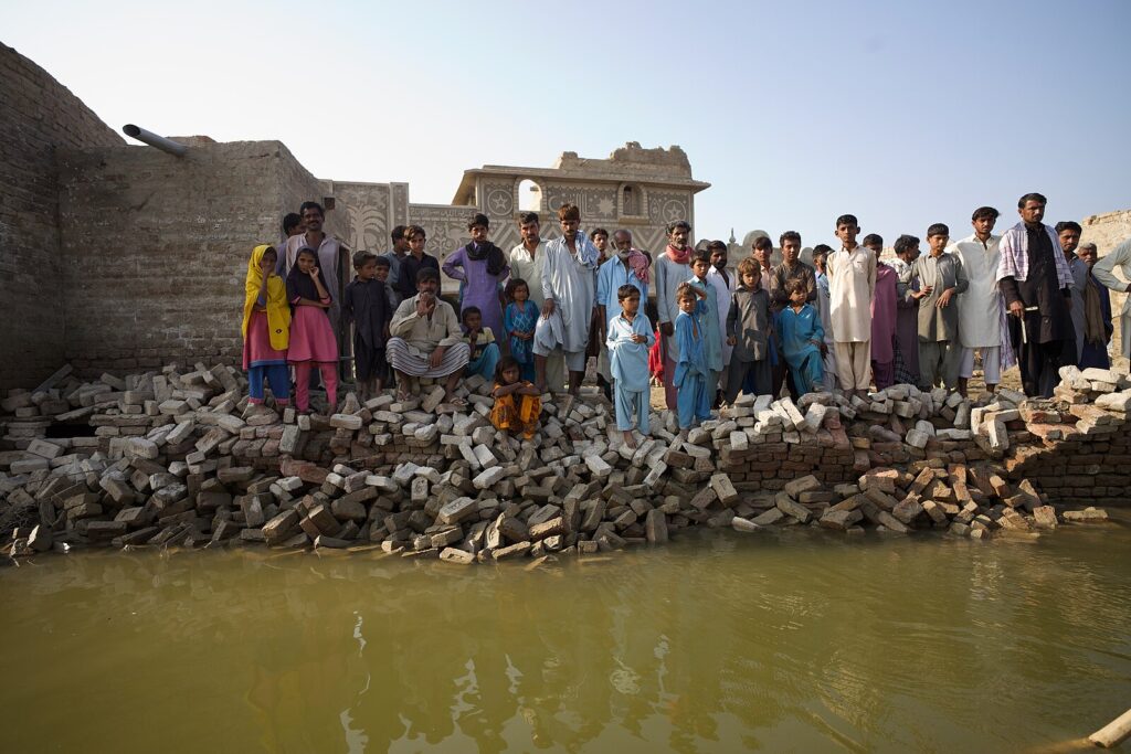 A crowd of people stand on a pile of bricks that crumble into the still flood waters in front of them.