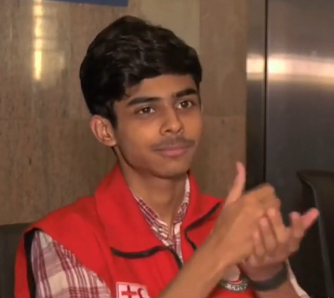 Kashaf Alvi has dark hair and is wearing a red vest over a white and red plaid shirt. He looks to the right while holdings his hands at chest height to sign.