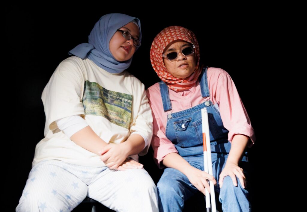 Two young Malaysians sit on a stage. One holds a white cane.