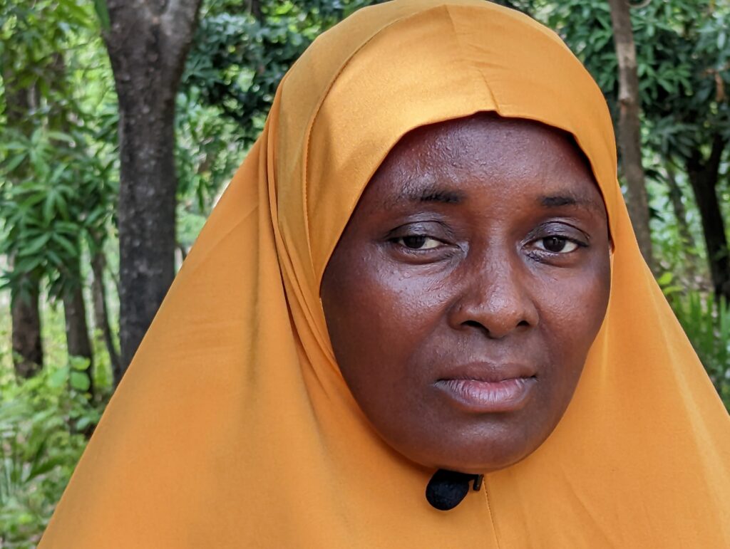 Amina Saleh, a woman wearing a mustard-yellow hijab, looks toward the camera while standing outdoors.