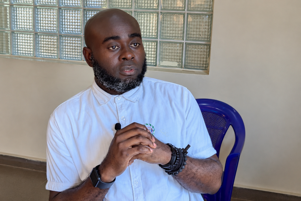 David Olusola, a Nigerian man with a shaved head and a salt-and-pepper beard, sits indoors wearing a white short-sleeved button-up shirt.