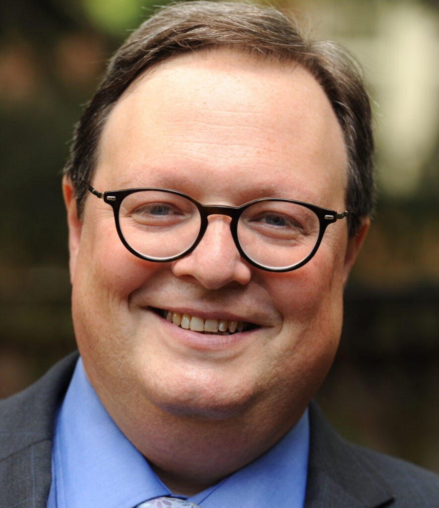 Eric Rosenthal smiling at the camera, wearing glasses, a dark suit, blue shirt and patterned tie.