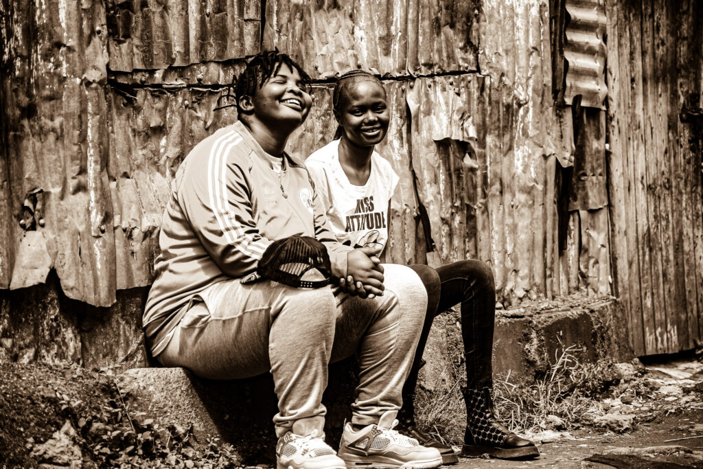Two queer friends laugh freely against the backdrop of a corrugated wall in Kenya.