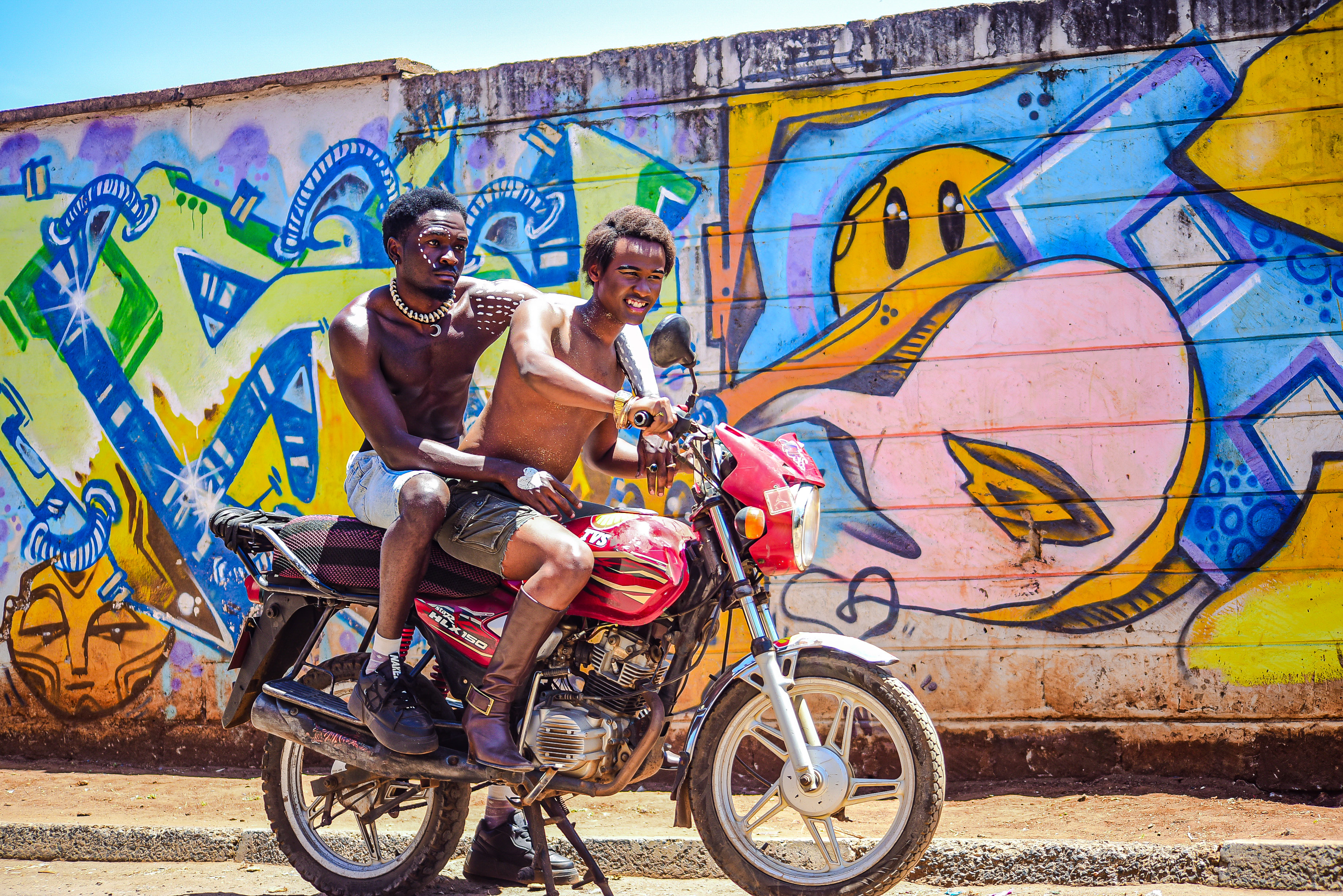Two African men sit on a red motorcycle in front of a brightly colored graffiti mural.